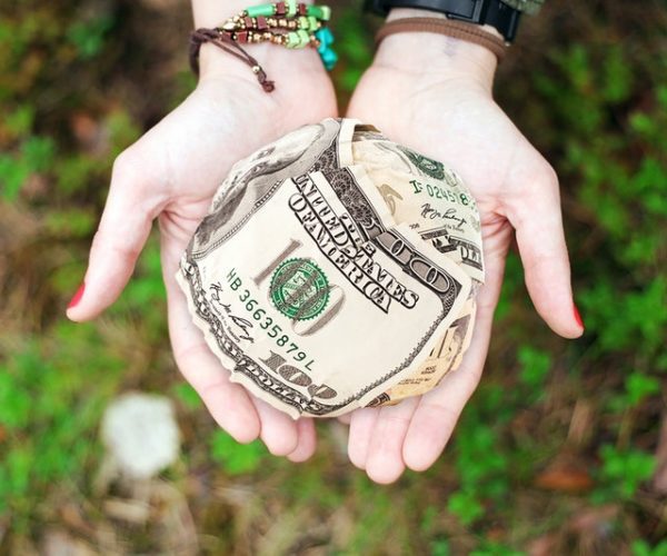 A pair of hands holding a crumpled ball of US dollar bills, symbolizing an opportunity to manifest money, with a focus on the currency and bracelets worn by the hands.
