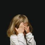 A young child with blonde hair symbolizing Inner Child Healing covers their face with both hands against a dark background, wearing a white shirt.