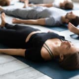 A group of adults lying on blue yoga mats in savasana pose, practicing breathwork therapy in a peaceful indoor setting.