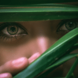 Close-up of a person's eyes peeking through green leaves, focusing on the detailed irises and eyelashes. The overall mood is mysterious and intimate.